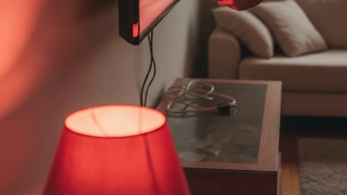 A person's hands plugging a streaming stick into the back of a wall-mounted television in a living room with red lamp light.