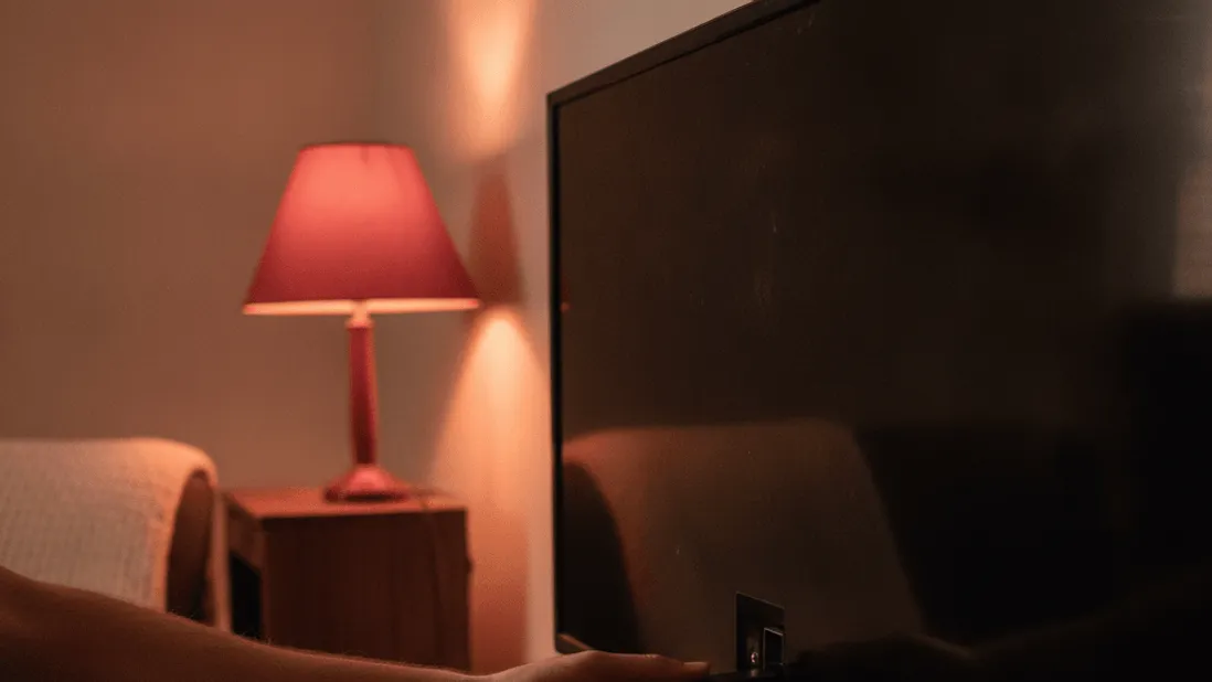 A person's hand inserts a small streaming stick into the back of a wall-mounted television, lit by warm lamp light.