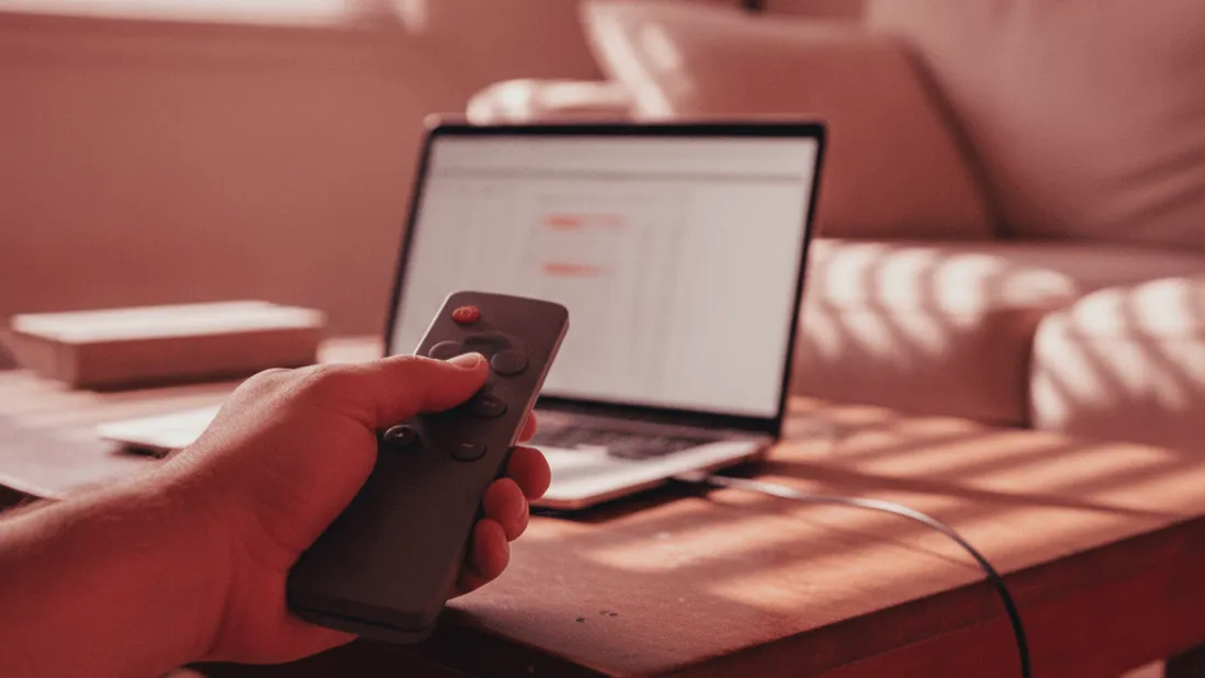 A hand holds a remote control over a coffee table with a laptop and a red cast from light.