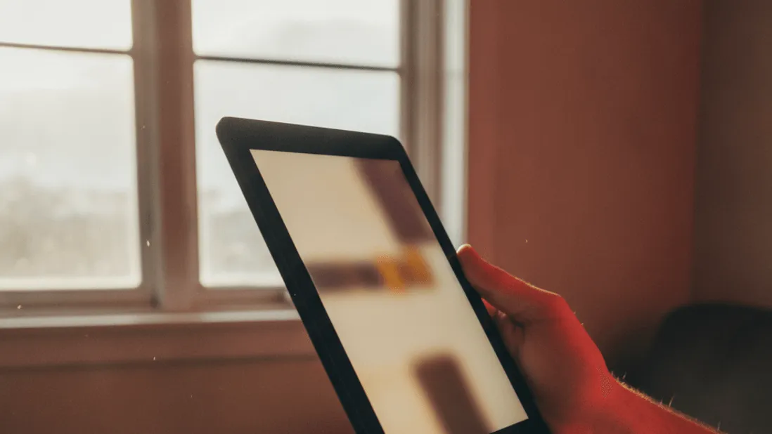 A hand holds a tablet near a window, a charging cable attached, with red light on the hand.