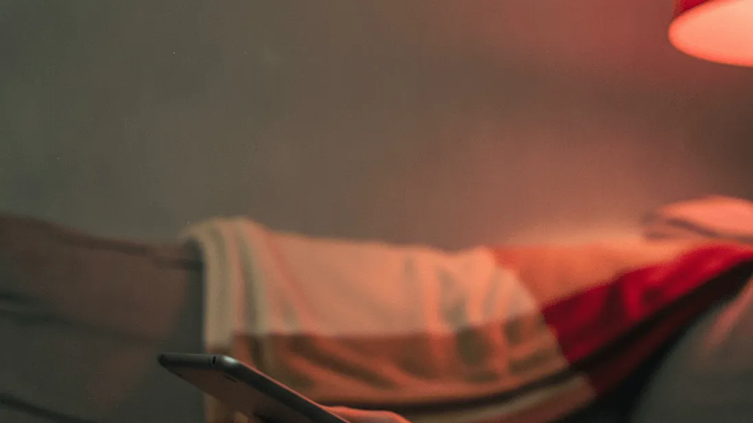 A hand holds a tablet on a grey sofa cushion with a red lamp glow and a blurred throw blanket.
