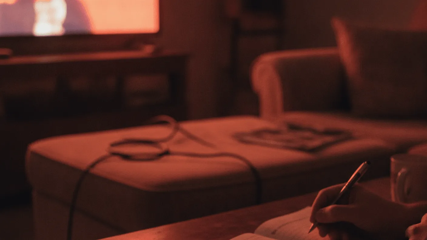 A remote control and notebook on a coffee table, with a person writing and a television in the background.