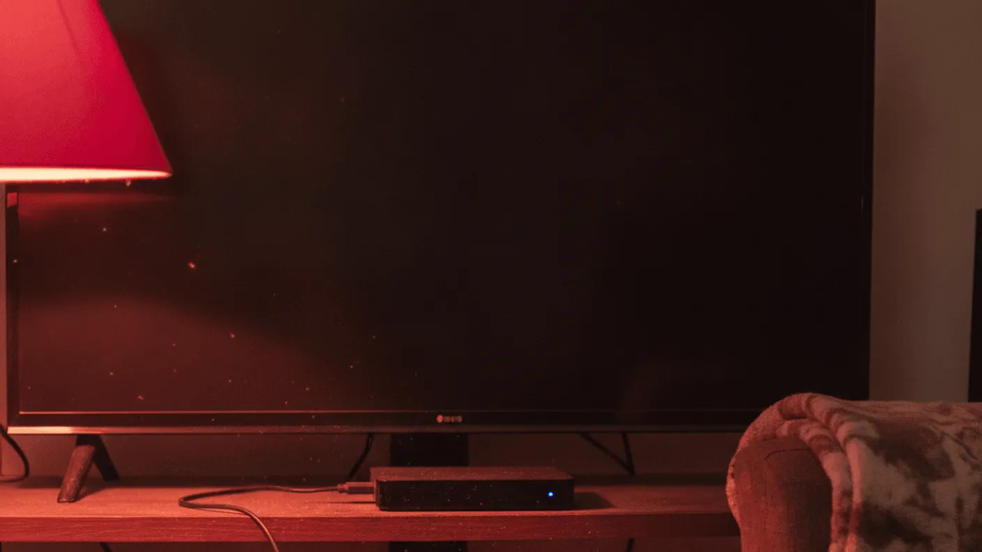 A small black media box sits on a wooden shelf under a switched-off television, with a red-shaded lamp to the left.