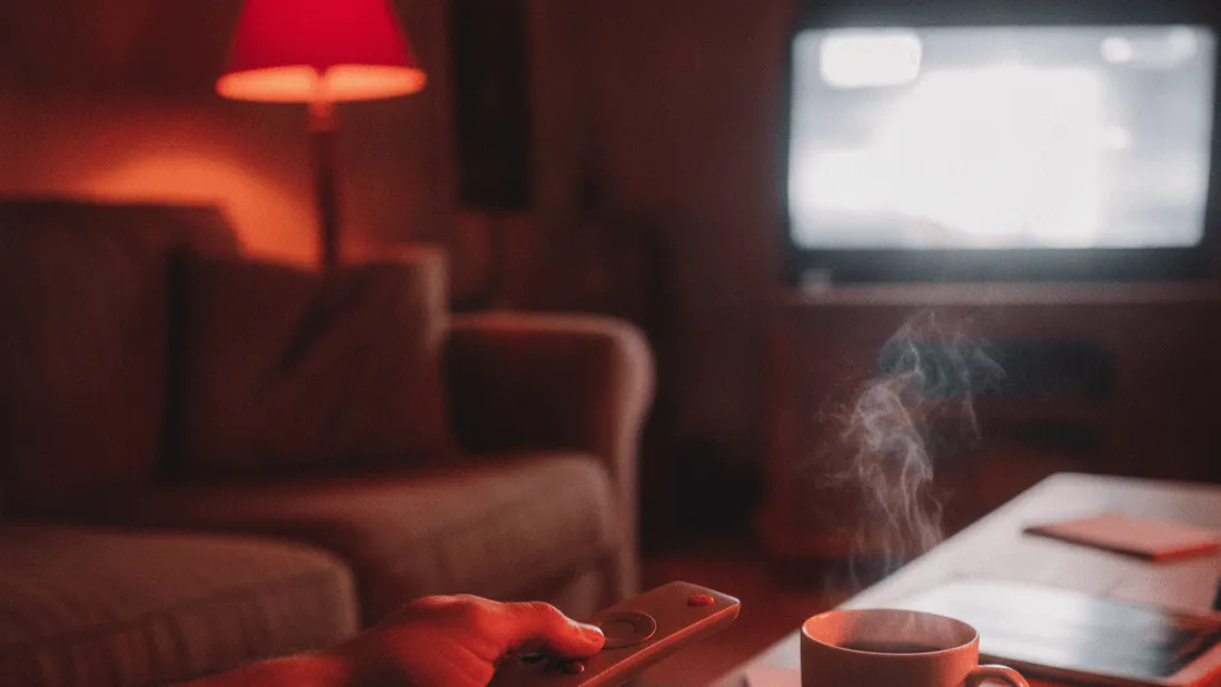 A hand holds a remote control above a coffee table with a steaming mug of tea at night.
