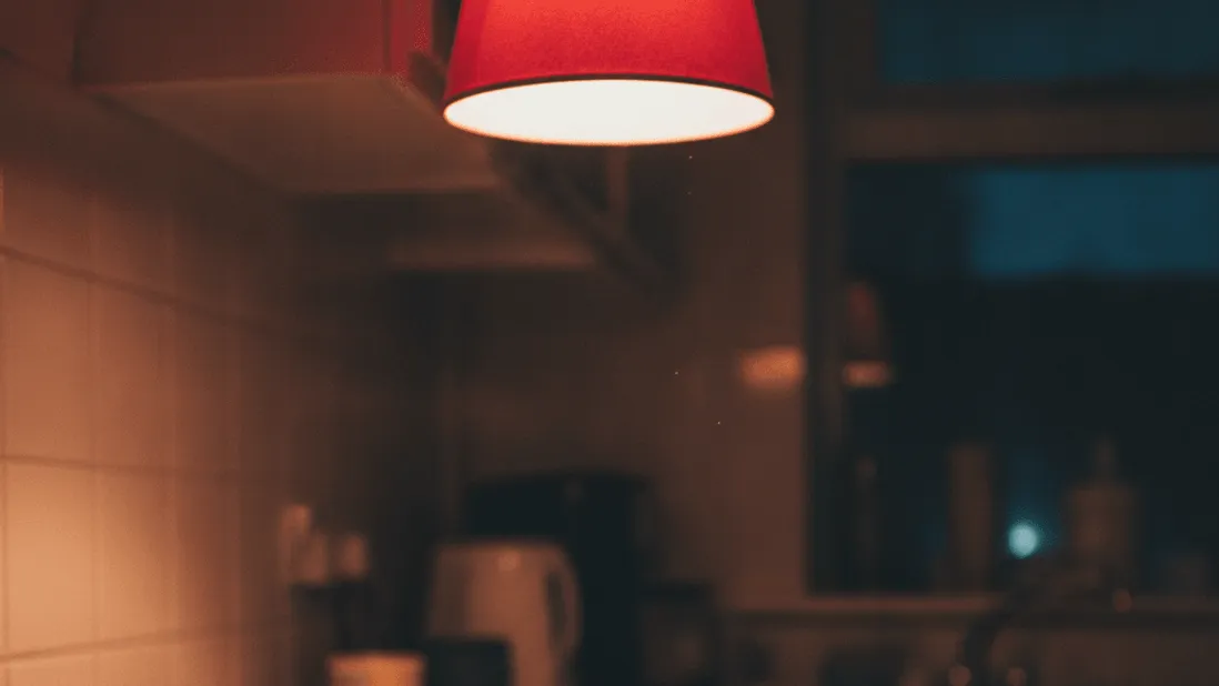 A man's hand rests on a smartphone charging on a kitchen counter at night, with a mug and a red lamp overhead.