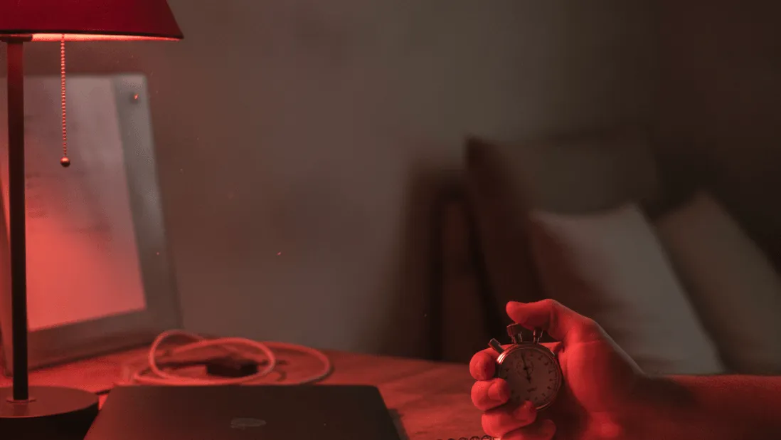A hand holds a stopwatch beside a laptop and a notebook on a desk under the red glow of a lamp.