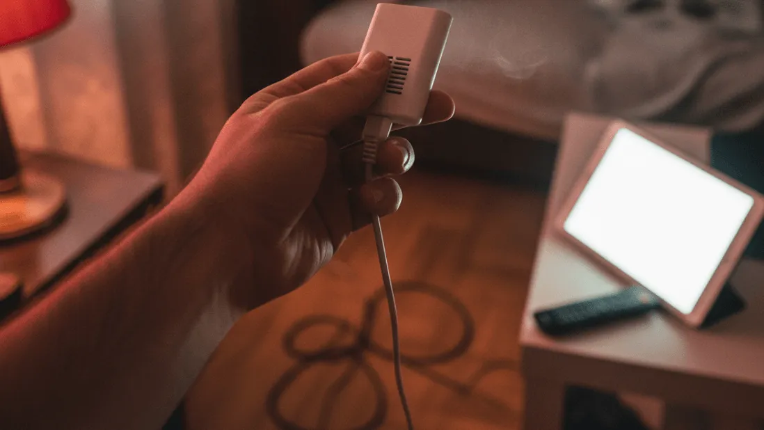 A hand holds a warm plastic dongle unplugged from a wall socket, with faint heat haze near its vents.