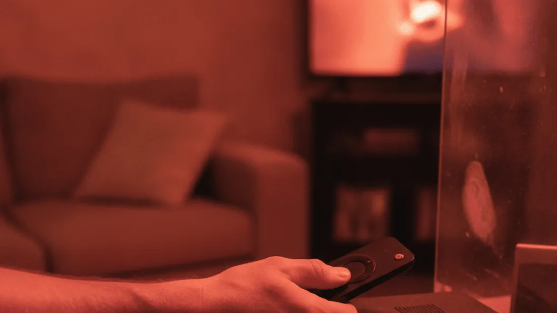 A hand holds a streaming remote near a set-top box on a shelf, lit by a red-tinged evening lamp.