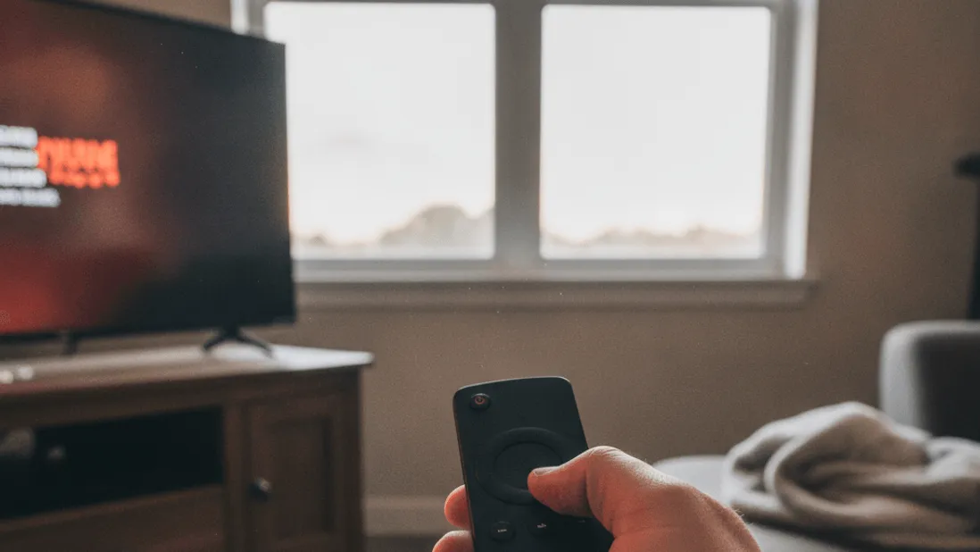 A close-up of a hand holding a remote, with a television, cables, and a window in the background.