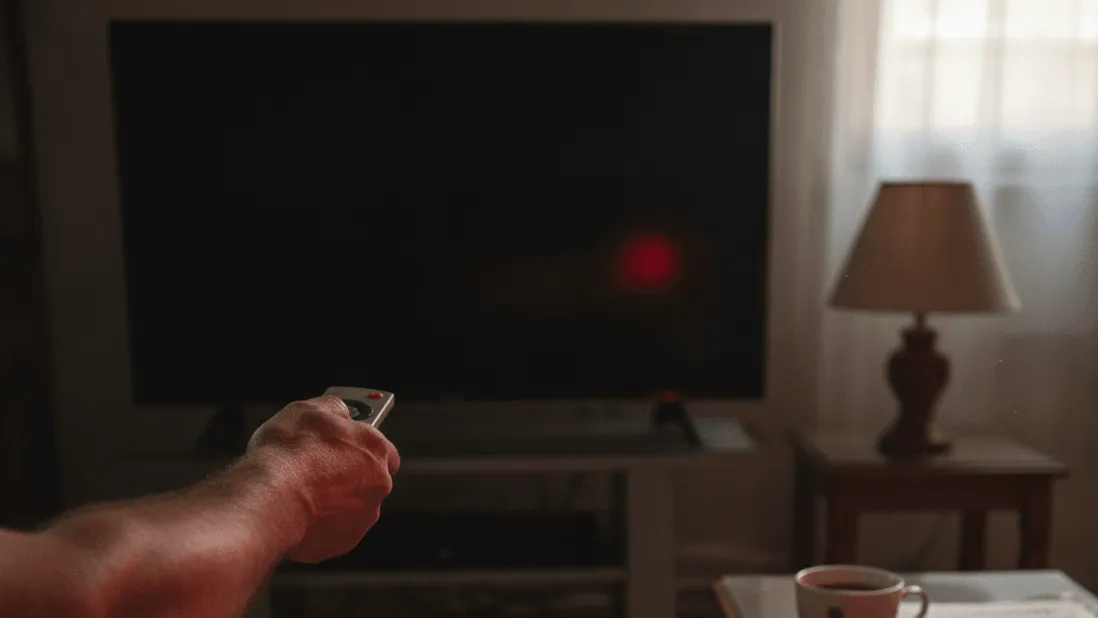 A hand holds a remote toward a dark television screen, with a cup of tea and newspaper on a side table.