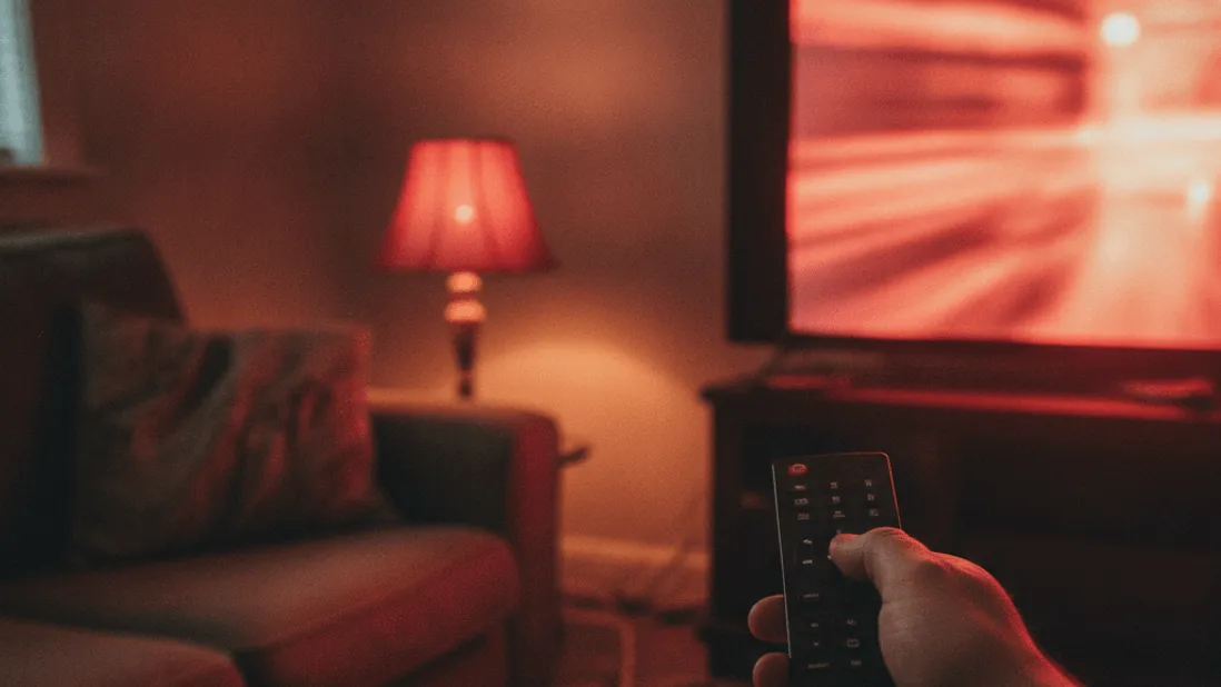 A hand holds a remote in a dimly lit living room with a red-tinged television screen and a notepad on the sofa.